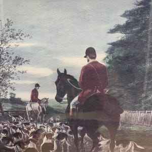 May include: A framed print depicting a fox hunting scene with two riders on horseback wearing red coats and black helmets. A pack of hounds in white, black, and brown markings runs across the foreground. The setting includes trees and a wooden fence.