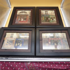 May include: A set of four square wall frames with black borders and dark brown inner mats. Each frame contains a photographic print of a European storefront with architectural details, including signs reading Antoinette, Cafe Bar, and Le Casserole.