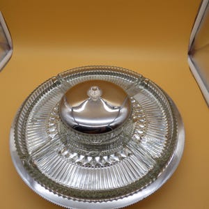 May include: A circular glass serving platter featuring four distinct partitioned sections with a ribbed texture. A central glass bowl with a polished chrome lid sits in the middle of the platter, all resting on a silver-toned metal base tray.
