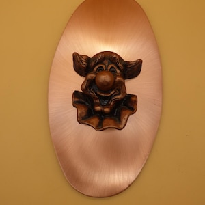 May include: A wall plaque featuring a central three-dimensional bronze-toned clown face with a ruffled collar, mounted on a smooth, oval-shaped copper-colored metal plate with a brushed finish and a distinct vertical grain pattern.