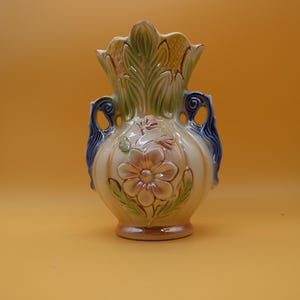 May include: A ceramic vase with a cream-colored body, featuring a raised floral relief design in pink and green. The vase has a scalloped rim with green leaf-like details and two dark blue, scrolled handles on the sides, finished with a glossy glaze.
