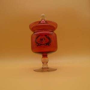May include: A translucent amber-colored glass candy dish with a matching lid and a pedestal base. The front of the glass container features a black printed graphic of a laurel wreath surrounding the text 40 Anniversary with a ribbon detail.