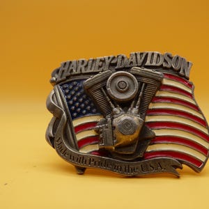 May include: A rectangular metal belt buckle featuring a raised silver-tone V-twin motorcycle engine centered over an American flag with red, white, and blue enamel. The text HARLEY-DAVIDSON is at the top, and Made with Pride in the U.S.A. is at the bottom.