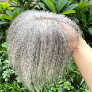 Puede incluir: Un postizo de cabello con hebras lisas de color gris plateado y una base visible de color piel en la coronilla. El cabello está peinado con una raya central, mostrando un área del cuero cabelludo de aspecto natural donde se fijan las fibras.