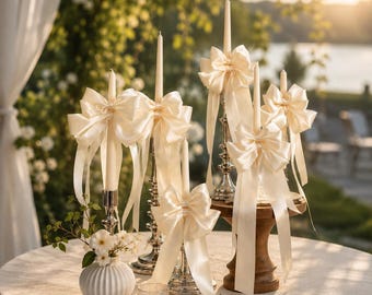 May include: A collection of five cream-colored taper candles, each adorned with a large, matching satin ribbon bow. The candles are held in a mix of silver-toned metal and wooden candle holders, arranged on a round table with a white cloth.
