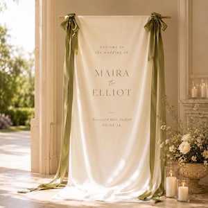 May include: A cream-colored fabric wedding welcome sign hanging from a horizontal rod, featuring dark text that reads "welcome to the wedding of MAIRA & ELLIOT Brutsyard Hall, Suffolk 09-03-24". Long olive green ribbons are tied at the top corners.