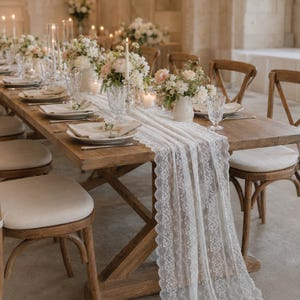 May include: A long white lace table runner draped over a rustic wooden dining table. The table is set with white ceramic plates, folded beige napkins, clear glass tumblers, and small white vases filled with white and light pink floral arrangements.