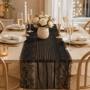 May include: A black lace table runner featuring a central striped pattern and scalloped floral edges, displayed on a light wood dining table with white plates, gold napkin rings, tall champagne flutes, and two lit taper candles.