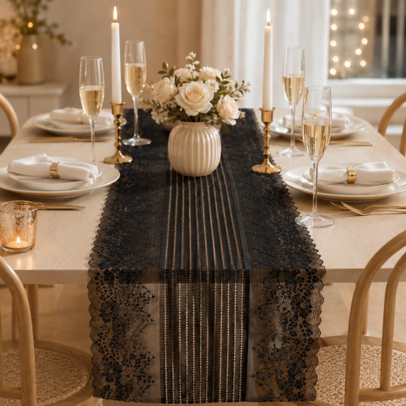 May include: A black lace table runner featuring a central striped pattern and scalloped floral edges, displayed on a light wood dining table with white plates, gold napkin rings, tall champagne flutes, and two lit taper candles.
