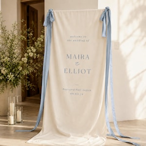 May include: A vertical cream-colored fabric banner featuring blue text that reads: welcome to the wedding of MAIRA & ELLIOT, Brutyard Hall, Suffolk, 09-03-24. The banner is accented with long, light blue satin ribbons tied at the top corners.