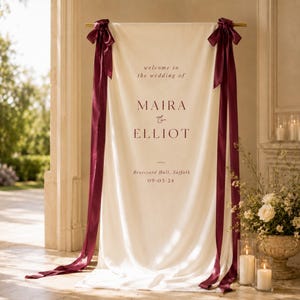 May include: A cream-colored fabric wedding welcome sign hanging from a gold rod, featuring dark red text that reads: welcome to the wedding of MAIRA & ELLIOT, Brutsyard Hall, Suffolk, 09-03-24. The banner is accented with long, dark red satin ribbons.