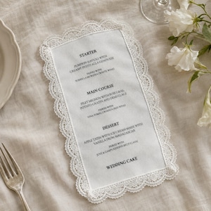 Könnte beinhalten: Eine rechteckige Menükarte aus weißem Stoff mit einer aufwendigen Spitzenbordüre. Der aufgedruckte Text listet eine Vorspeise aus Kürbis-Ravioli, ein Hauptgericht aus Filet Mignon mit Foie Gras und Kartoffelgratin, ein Dessert sowie eine Hochzeitstorte auf.
