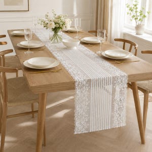 May include: A white lace table runner with a floral pattern and vertical stripes draped over a light wood dining table. The table is set with white ceramic plates, clear glass stemware, and a glass vase filled with white flowers.