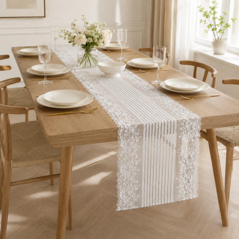 May include: A white lace table runner with a floral pattern and vertical stripes draped over a light wood dining table. The table is set with white ceramic plates, clear glass stemware, and a glass vase filled with white flowers.