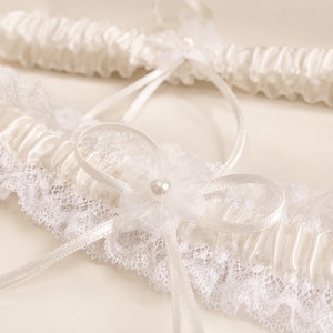 May include: A pair of white wedding garters featuring gathered satin fabric and delicate lace trim. Each garter is adorned with a sheer organza flower centered with a small faux pearl and accented by thin satin ribbon loops and long trailing ribbons.