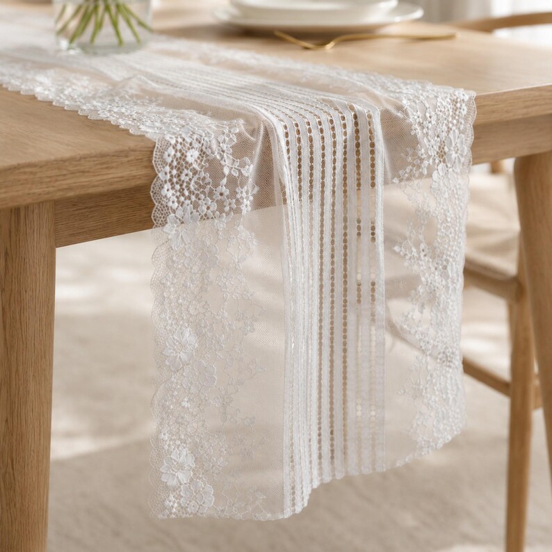 May include: A white table runner featuring a sheer mesh center panel with vertical stripes and wide, intricate floral lace borders along the edges. The fabric is lightweight and semi-transparent, draping over the side of a wooden table surface.
