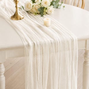 May include: A sheer, off-white fabric table runner draped over a white table. The runner is accented by a gold-toned metal candle holder, a small white pillar candle, and a bouquet of white roses with green eucalyptus leaves.