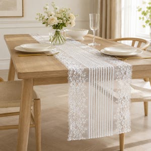May include: A white lace table runner featuring a central panel of vertical stripes and dotted lines, flanked by intricate floral lace borders. The runner is draped over a light-toned wooden dining table set with white plates, a glass, and a vase.