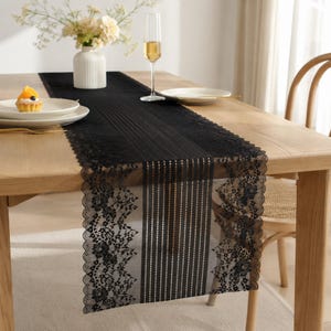 May include: A black lace table runner featuring a central panel of vertical stripes flanked by intricate floral patterns and scalloped edges, draped over a light wood dining table with a white vase, a champagne flute, and a small fruit tart.