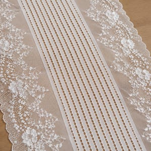 May include: A white lace table runner featuring a central panel of vertical stripes and openwork detailing, flanked by wide borders of intricate floral embroidery and scalloped edges. The fabric is sheer and lightweight with a delicate texture.