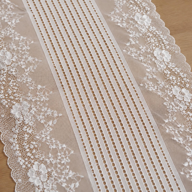 May include: A white lace table runner featuring a central panel of vertical stripes and openwork detailing, flanked by wide borders of intricate floral embroidery and scalloped edges. The fabric is sheer and lightweight with a delicate texture.
