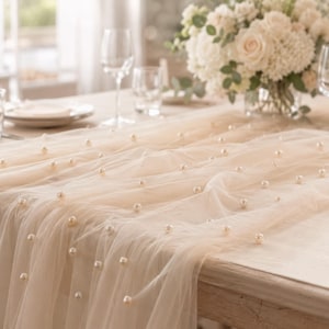 Peut inclure: Un chemin de table en tulle transparent de couleur champagne, orné de petites perles artificielles crème, rondes et régulièrement espacées. Le tissu léger retombe sur le bord de la table, créant des plis doux et un aspect texturé.