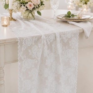 Puede incluir: Un camino de mesa de encaje blanco con un patrón floral repetitivo de rosas y bordes festoneados. La tela es semitransparente y cuelga sobre el borde de una mesa, mostrando detalles de bordado intrincados a lo largo de toda la pieza.