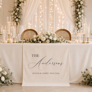 May include: A cream-colored fabric table runner featuring black printed text that reads THE Andersons, OLIVIA & JAMES | 09.05.2026. The runner is draped over a table decorated with a white floral centerpiece, tall glass candle holders, and two chairs.