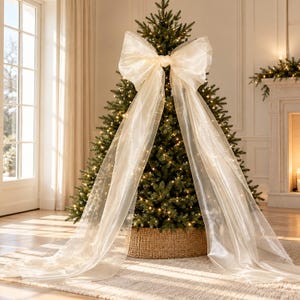Peut inclure: Un grand nœud en organza transparent blanc cassé avec deux longs pans fluides drapés sur le devant d'un sapin de Noël vert. L'arbre est illuminé par des guirlandes lumineuses blanc chaud et repose dans une base en panier tressé cylindrique.