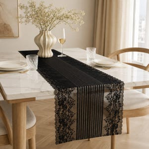 May include: A black lace table runner featuring a central panel of vertical stripes flanked by intricate floral lace borders with scalloped edges, draped across a white marble dining table with place settings and a cream-colored vase.