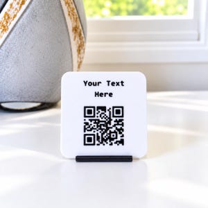 Peut inclure: Un panneau carré blanc aux coins arrondis maintenu par un support noir. Le panneau présente le texte imprimé en noir "Your Text Here" au-dessus d'un code QR noir et blanc. Le design est épuré et simple, offrant un affichage clair.