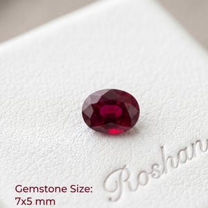 May include: An oval-cut, deep red ruby gemstone with a faceted surface. The stone is positioned on a white textured surface with the text "Roshan" embossed in the corner and the printed text "Gemstone Size: 7x5 mm" visible in the lower left.