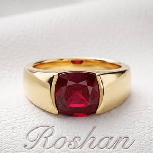 Op de afbeelding: Een gouden ring met een dieprode edelsteen in kussenslijpvorm in het midden. De ringband is glad en gepolijst met een breed, solide ontwerp. Het woord Roshan is in cursief schrift onder de ring op het gestructureerde witte oppervlak gedrukt.