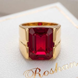 Könnte beinhalten: Ein Goldring mit einem großen, rechteckigen, im Smaragdschliff geschliffenen tiefroten Edelstein, der in ein poliertes Goldband gefasst ist. Der Ring liegt auf einer weißen, strukturierten Oberfläche mit dem Schriftzug Roshan in Gold.