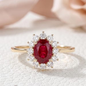 May include: A gold ring featuring a central oval-cut deep red ruby gemstone surrounded by a halo of twelve round brilliant-cut white diamonds. The band is polished yellow gold, and the setting holds the stones in a classic floral-inspired arrangement.