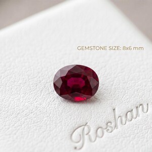Könnte beinhalten: Ein oval geschliffener, tiefroter Rubin-Edelstein mit facettierter Oberfläche. Der Stein liegt auf einer weißen, strukturierten Oberfläche mit dem Text GEMSTONE SIZE: 8x6 mm und dem Wort Roshan in der unteren rechten Ecke.