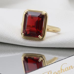 Puede incluir: Un anillo de oro con una gran piedra preciosa de color rojo intenso, corte esmeralda rectangular, engastada en un bisel de oro pulido y liso con cuatro pequeñas garras en las esquinas. El anillo está sobre una superficie blanca y se refleja.
