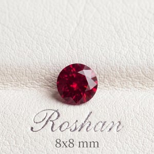 Puede incluir: Una piedra preciosa redonda, facetada y de color rojo intenso con talla brillante. La piedra se sitúa sobre el texto "Roshan" y la medida "8x8 mm" impresos en una elegante fuente cursiva sobre una superficie texturizada de color blanco roto.