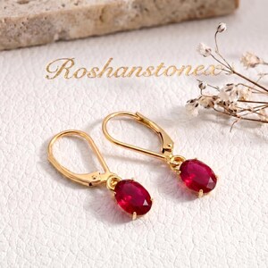 Puede incluir: Un par de pendientes dorados con cierre de palanca y piedras de color rojo intenso de corte ovalado. El texto 'Roshanstones' es visible sobre las joyas. Los pendientes tienen un acabado de metal pulido y un mecanismo de cierre articulado.
