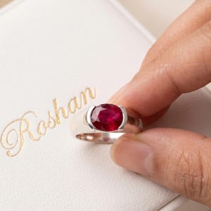 May include: A silver-toned ring featuring a large, oval-cut, deep red gemstone set in a tension-style mounting. The ring is positioned against a white textured surface that displays the gold-coloured cursive text Roshan printed on the left side.