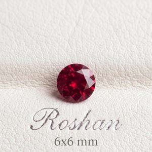 Può includere: Una gemma rotonda, sfaccettata e di colore rosso intenso che misura 6x6 mm. La pietra è posizionata sopra il testo "Roshan" e le dimensioni "6x6 mm" stampate in un elegante carattere corsivo scuro su una superficie testurizzata color bianco sporco.
