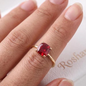 Op de afbeelding: Een gouden ring met een dieprode edelsteen in kussenslijpvorm, gezet in een zetting met vier pootjes. De ringband is dun en gepolijst en toont een levendige, gefacetteerde rode steen die op de vinger ligt en het licht reflecteert.