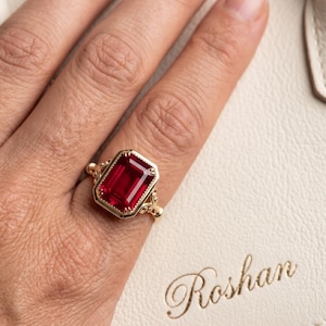 Peut inclure: Une bague en or ornée d'une grande pierre précieuse rouge foncé de taille émeraude rectangulaire, montée sur une monture en métal décorative avec des détails en filigrane sur l'anneau. La bague est portée avec l'inscription Roshan visible.