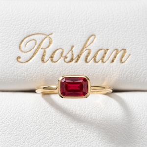 Può includere: Un anello in oro con una pietra preziosa rossa rettangolare incastonata a lunetta. L'anello è posizionato davanti a una superficie bianca testurizzata con la parola Roshan impressa in lettere dorate in corsivo sopra il gioiello.