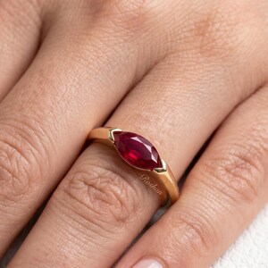 Op de afbeelding: Een gouden ring met een dieprode robijn in marquise-slijpvorm, gezet in een gladde metalen band. Het woord Roshan is in cursief schrift gegraveerd aan de zijkant van de gouden band, die de gefacetteerde steen vasthoudt in een gladde zetting.