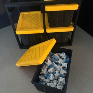 May include: A black storage rack holding three small black bins with bright yellow lids featuring a diamond grid pattern. A fourth matching black bin sits in front, filled with numerous white and blue mechanical keyboard switches.