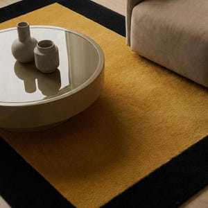 Peut inclure: Un tapis rectangulaire présentant un grand panneau central jaune moutarde encadré par une large bordure noire. Une table basse ronde et basse, au fini beige brillant et au plateau réfléchissant, porte deux petits vases en céramique gris clair.