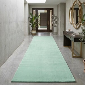 Peut inclure: Un long tapis de couloir rectangulaire d'une couleur vert menthe douce et unie. Le tapis présente une texture dense à poils ras et est placé au centre sur un sol en béton gris clair lisse, s'étendant du premier plan vers le fond de l'espace.