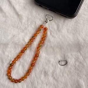 Peut inclure: Une dragonne de téléphone composée de perles rondes et translucides orange et vertes. La boucle de perles est reliée à un anneau métallique argenté et à une languette en plastique transparent, conçue pour un smartphone noir visible en haut.