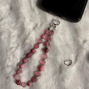 Peut inclure: Une dragonne de téléphone composée d'une boucle de perles rondes translucides roses et magenta, séparées par de petites entretoises en métal argenté. La dragonne est fixée à un smartphone noir par une languette en plastique transparent.